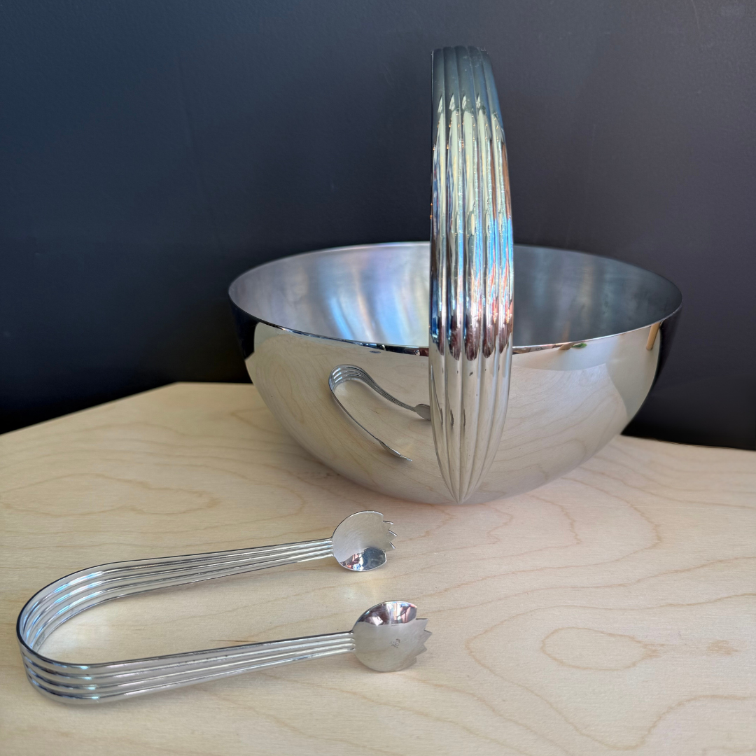 Set of silver kitchen utensils including a bowl, tongs, and measuring spoons on a wooden surface with a dark background.