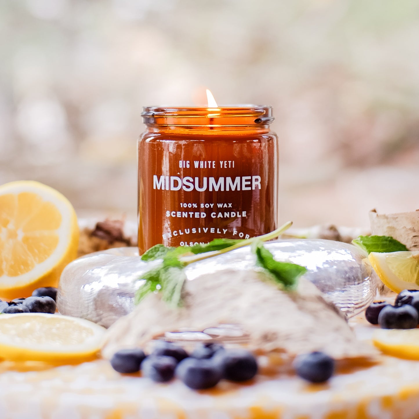 A soy wax candle with the label 'Midsummer' burning on a surface with fruits and herbs around it, scented of lemon, blueberry, mint, rosemary & birch