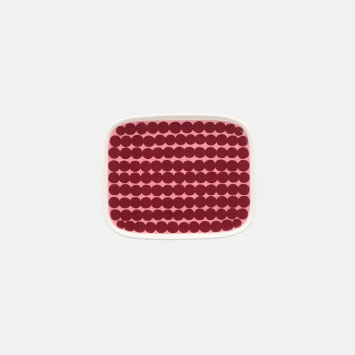 A square ceramic plate with rounded corners, featuring a repeating pattern of small red circles on a pink background, centered on a light gray studio backdrop.