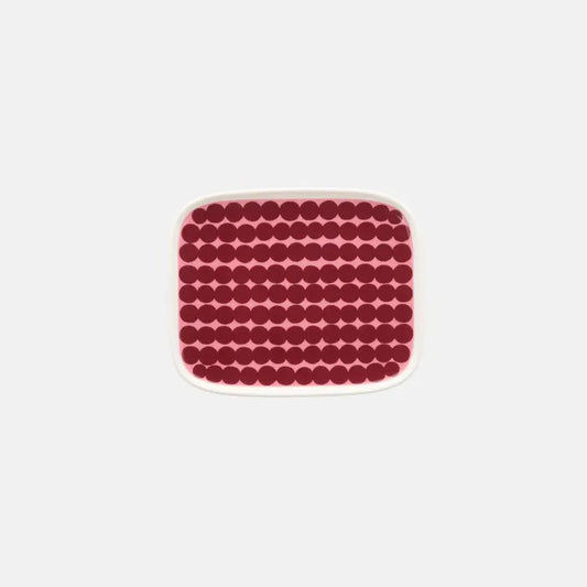 A square ceramic plate with rounded corners, featuring a repeating pattern of small red circles on a pink background, centered on a light gray studio backdrop.
