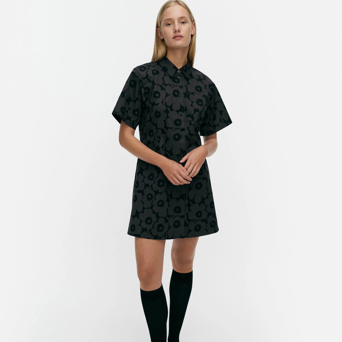 A model stands against a light gray studio background wearing a short-sleeve black collared dress with a tonal floral pattern, paired with black knee-high socks and black shoes.