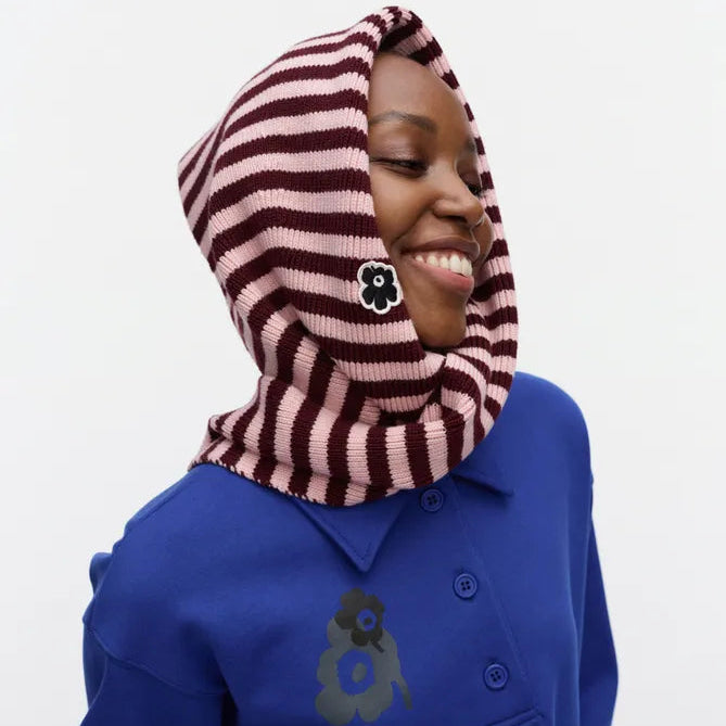 A smiling woman wearing a pink and dark red striped knit scarf with a flower-shaped decor, paired with a royal blue long-sleeve polo shirt against a plain white background.