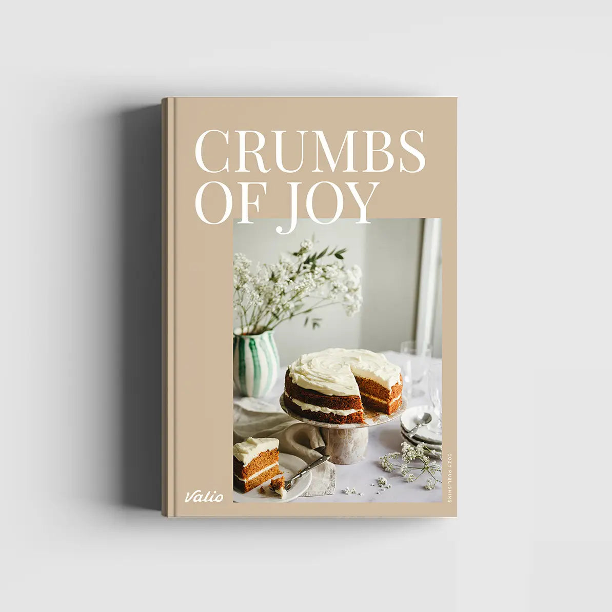 A hardcover book titled 'CRUMBS OF JOY' by Valio, featuring a warm beige cover with white text and a central photograph of a layered cake on a table, shown against a light gray background.