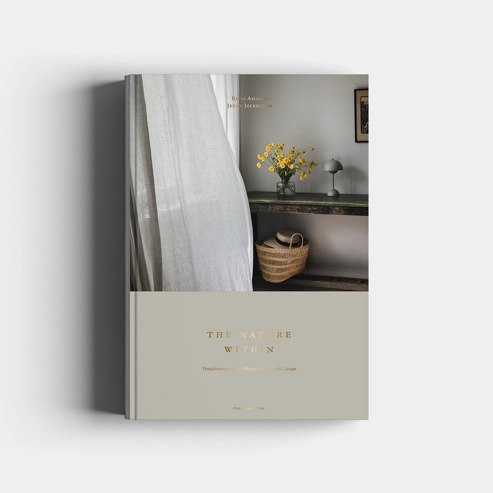 A hardcover book titled 'THE NATURE WITHIN' with a minimalist beige cover, featuring a photograph of a cozy interior scene on the upper half and gold foil-style title text on the lower half.