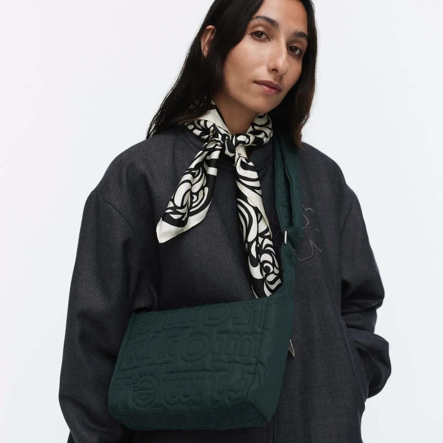 Person wearing a dark gray outfit with a green bag and a patterned scarf on a white background