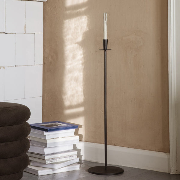 Tall black candle holder with dripping candle. There is a beige wall and a stack of books on the ground.