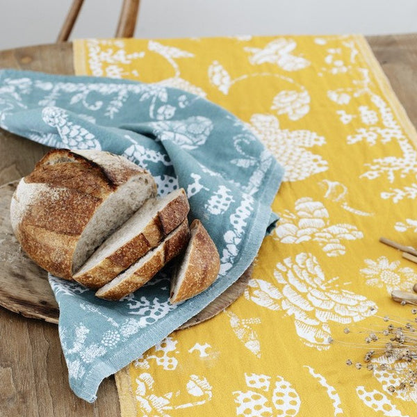 This linen tea towel has a flower and butterfly pattern. It is on a wooden table with a runner, dried flowers, and a loaf of bread.