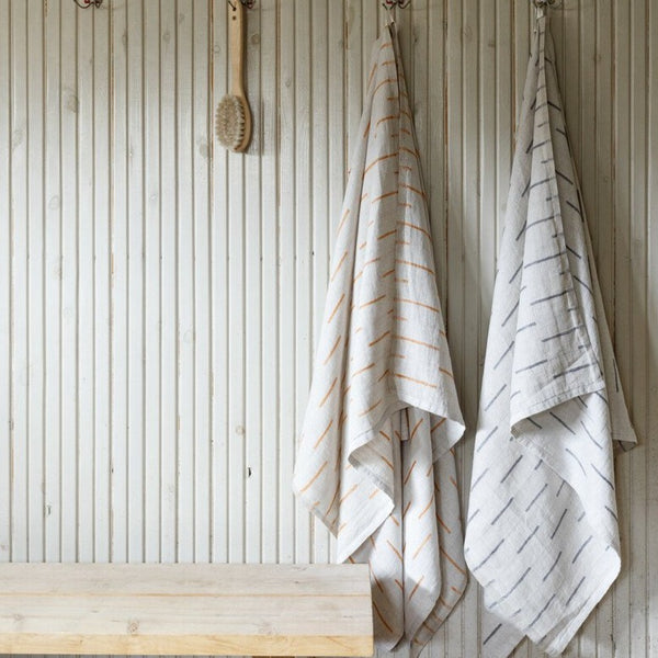 A white and gold towel, white and green towel, and scrubbing brush hanging from hooks against a wood slat wall. 