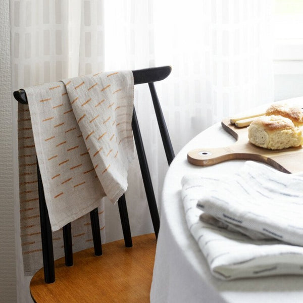 Linen towel draped over the back of a dining chair. Table covered in cloth with folded linens, a cutting board, and loaf of bread. 