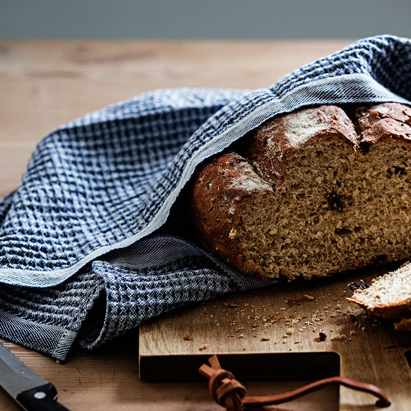 Loaf of bread wrapped in blue kitchen towel on cutting board. 