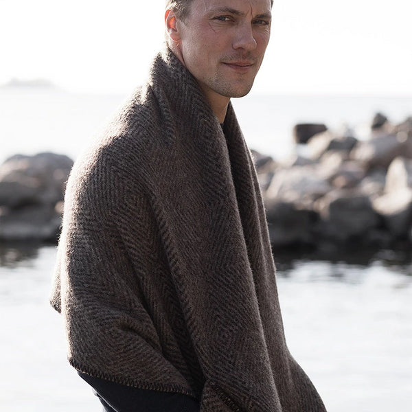 Man wrapped in wool shawl by rocks and water.