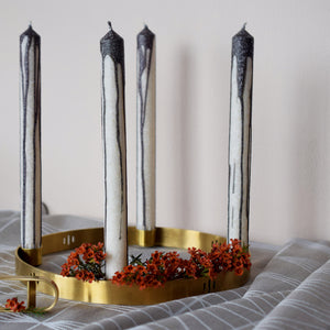 A round brass candle holder is holding 4 grey and white dipped candles. There is a beige tablecloth and red flowers.